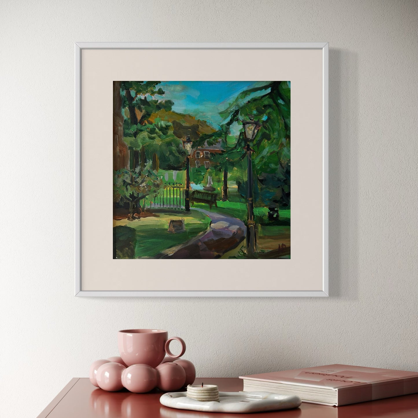 Framed painting of a park scene on a wall above a table with decor items. The painting is Chronicles of Malvern: Lantern Light original oil painting of the Narnia style lanterns in Malvern.