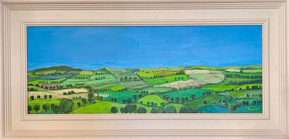A framed landscape painting depicting rolling green Cotswolds hills, fields, and trees under a bright blue sky, housed in a light beige wooden frame.