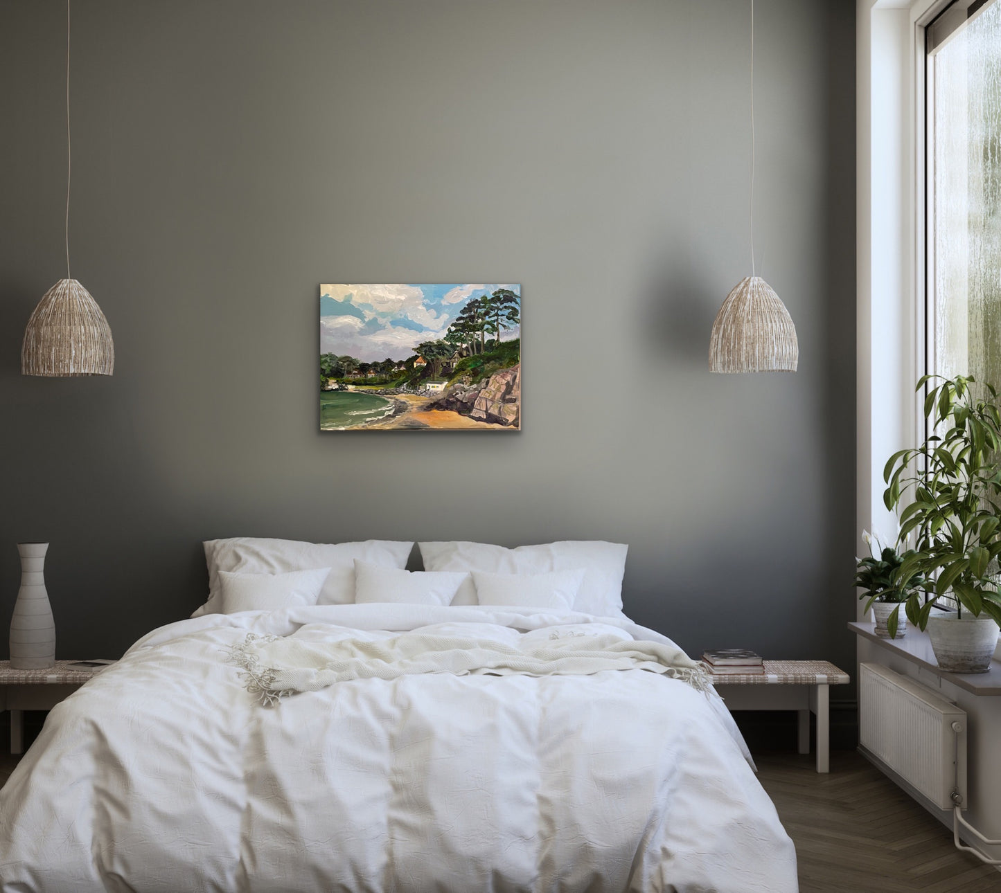 Painting of Caswell Bay in the Gower, Wales with white clouds in a blue sky and rocks and sea in foreground with trees and houses on the headland. Displayed on a grey wall with white bed in foreground.