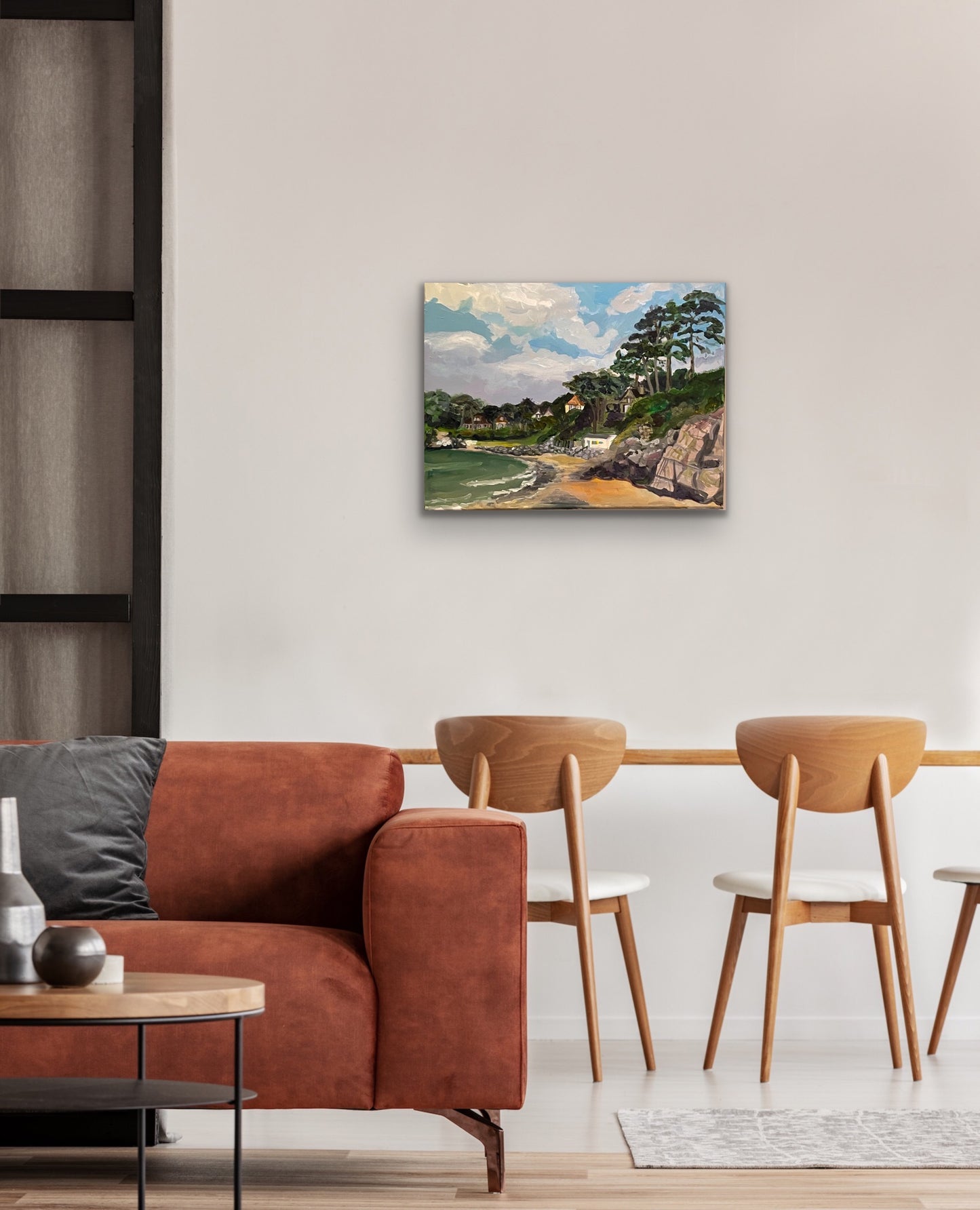 Painting of Caswell Bay in the Gower, Wales with white clouds in a blue sky and rocks and sea in foreground with trees and houses on the headland. Displayed against a white wall with pink sofa and wooden chairs in foreground.