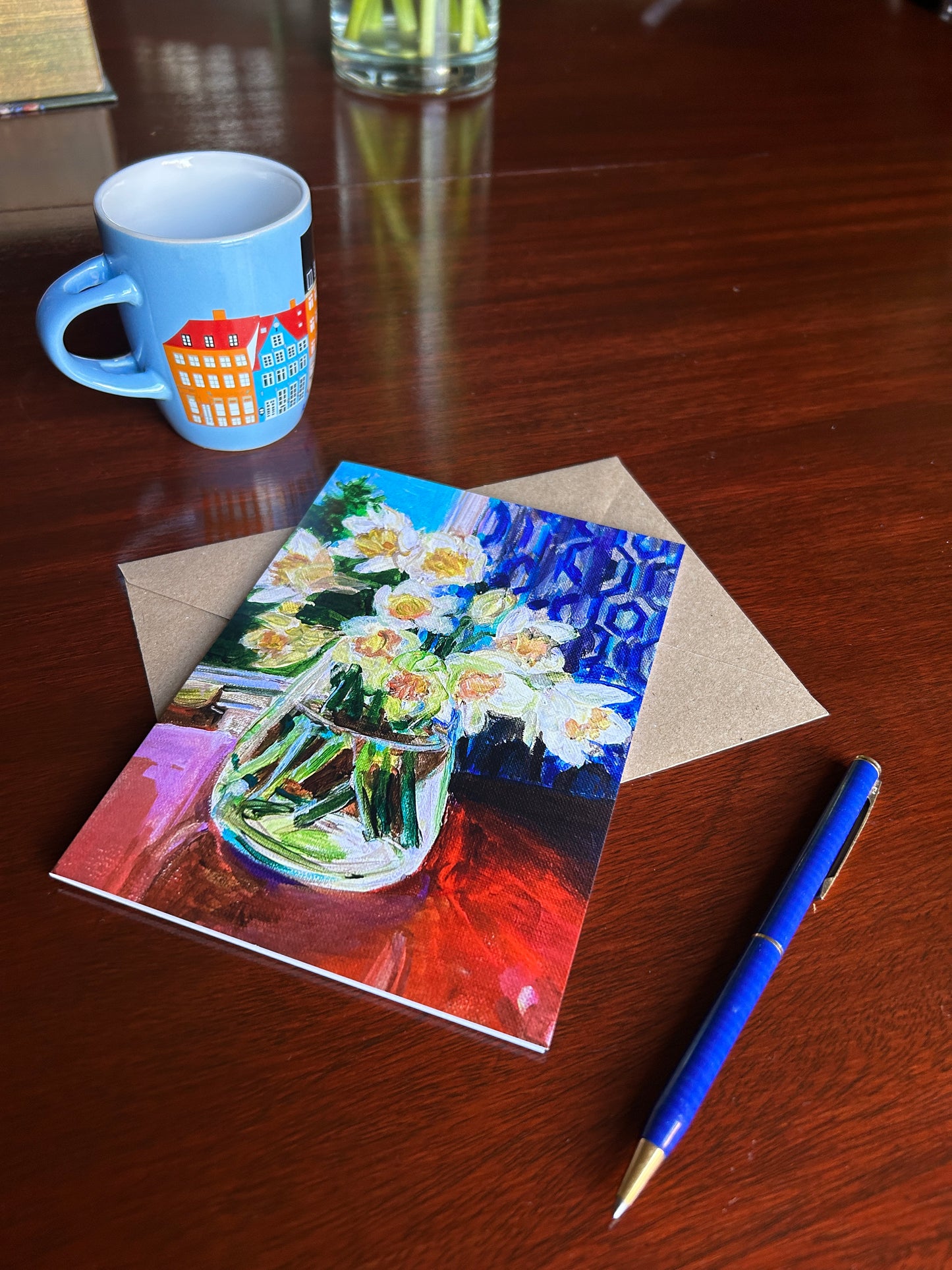 Greetings card of daffodils in a glass vase. Displayed with brown envelope on a wooden table with pen beside and mug behind.