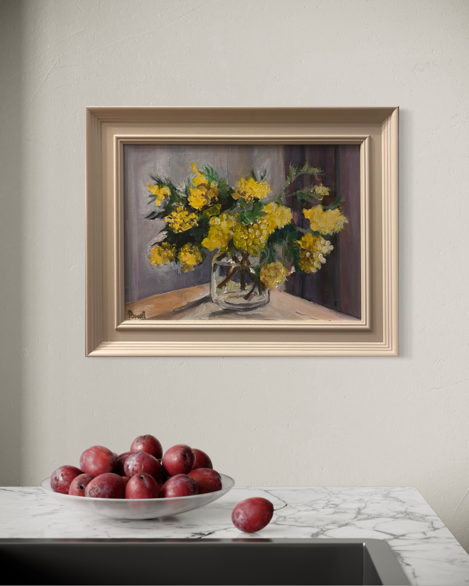 A framed still life painting of yellow flowers in a glass vase, displayed on a light beige wall above a marble countertop with a bowl of red plums.