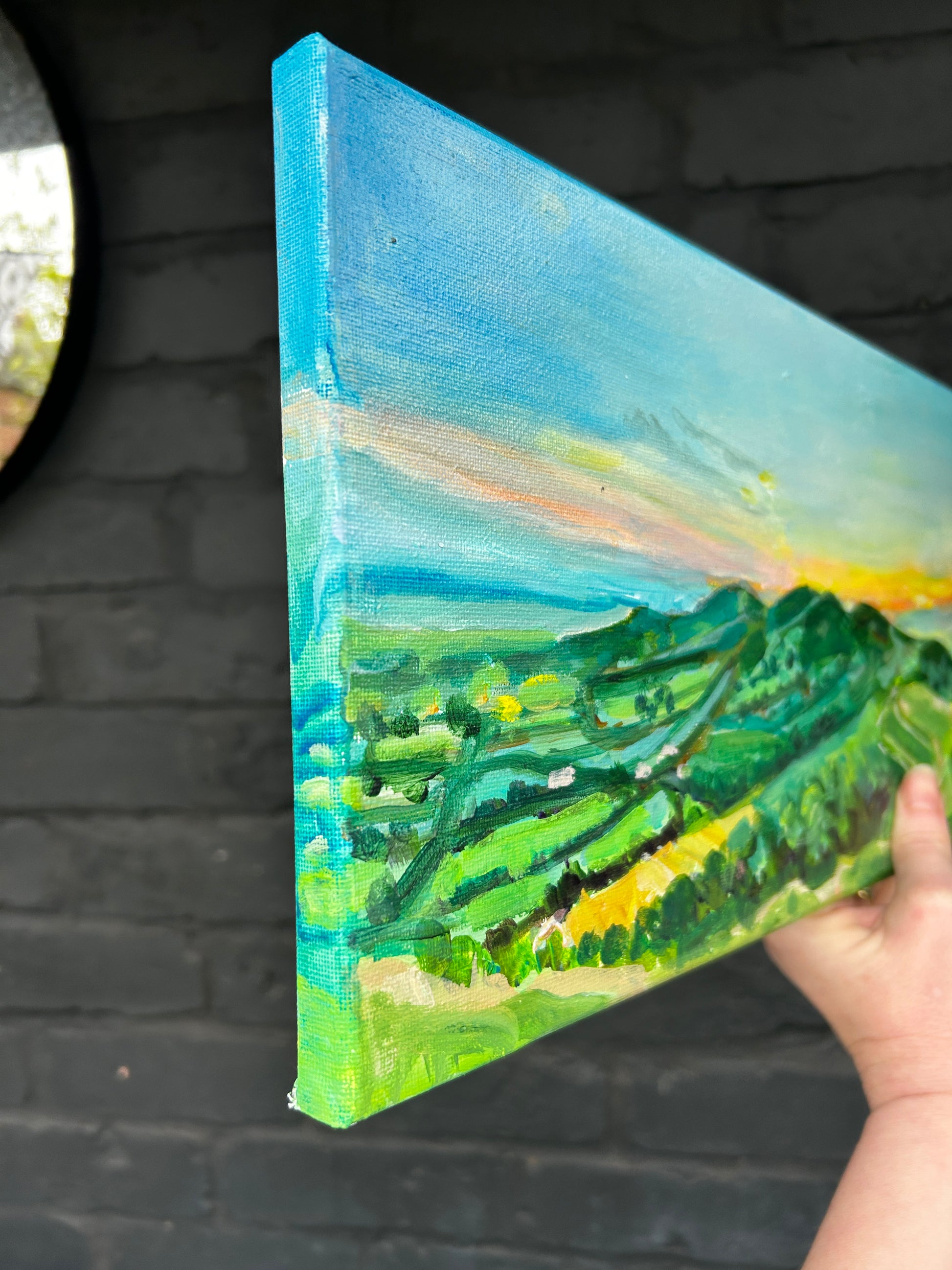 Original painting of Dawn at the Malvern Hills by Worcestershire Artist Margaret Powell canvas held in hand against black brick wall