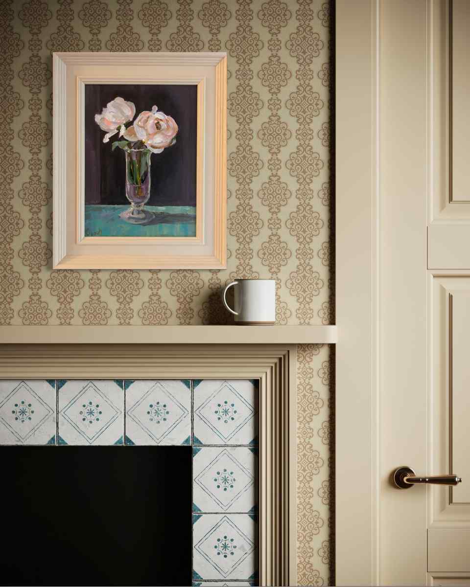 A framed still life painting of pink flowers in a glass vase, hung on a patterned wallpaper wall above a fireplace mantel with a white mug resting on it.