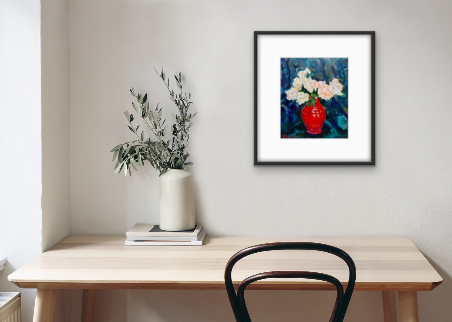 Framed print of a fine art painting of Blush Roses in a Red Vintage Vase. Displayed against a white wall with wooden table and chair in front and white vase containing leaves on the table. 