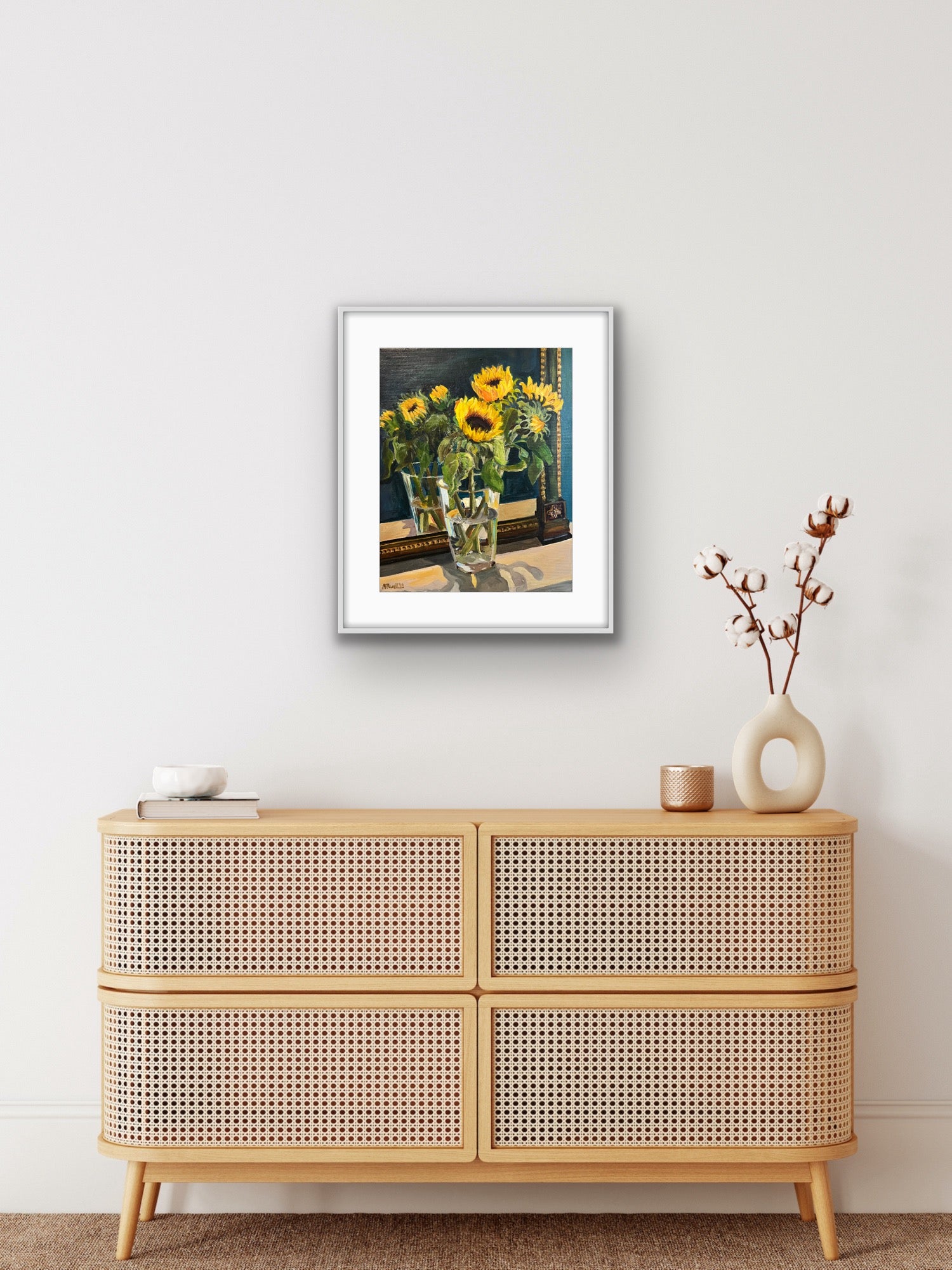 Print of a fine art painting of sunflowers displayed in a vase in front of a vintage mirror. Print framed in a white frame and displayed on a white wall above a beige wood cabinet. 